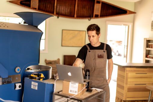 Man in an apron standing at a laptop that is connected to a machine that processes coffee.
