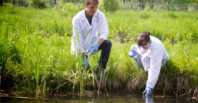 Two people in white lab coats take a water sample from a creek