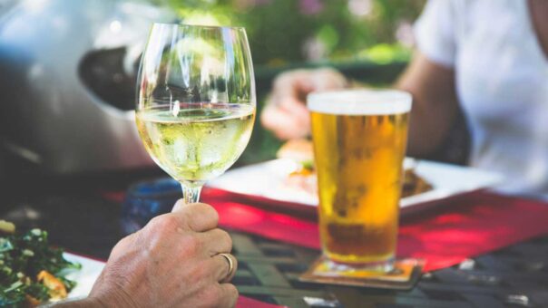 A close-up of a hand holding a glass of white wine with a beer on a table in the background