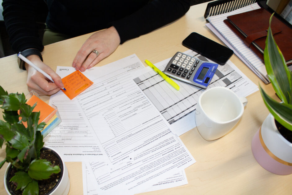 Person writing on a sticky note on a desk covered in tax related worksheets with a calculator, a highlighter, a mug and a phone