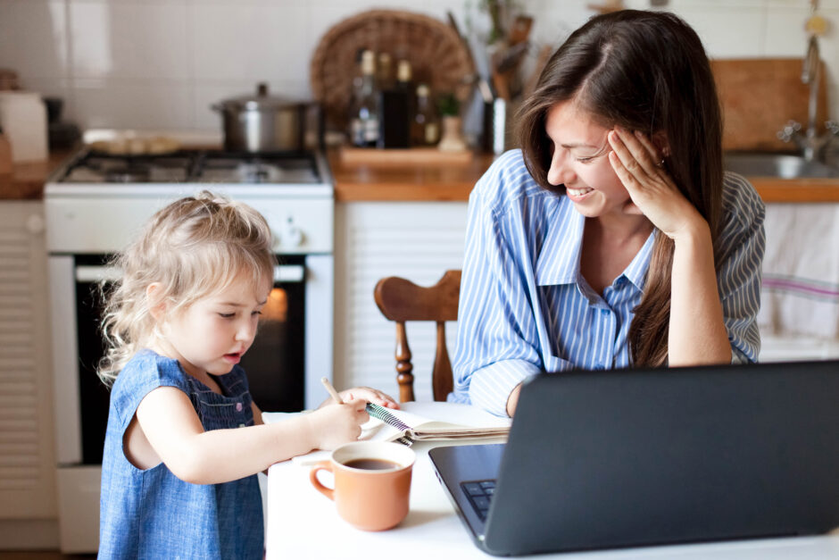 Work from home office with kid. Working mother using laptop. Cozy freelance workplace at kitchen table. Happy female business, woman career. Cute child drawing in mom notebook.