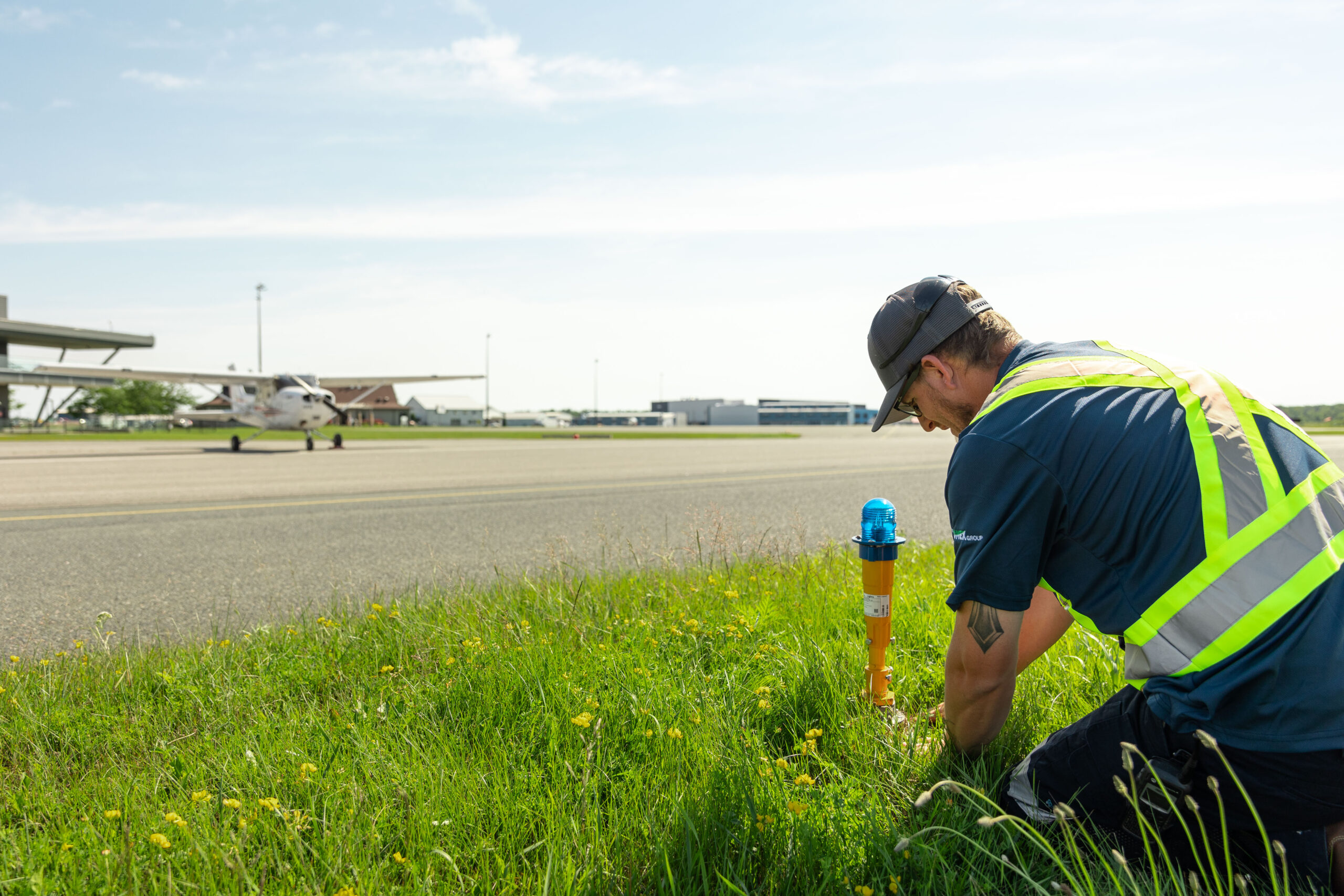 Person kneeling in grass working with an airport tarmac in the background with a plane